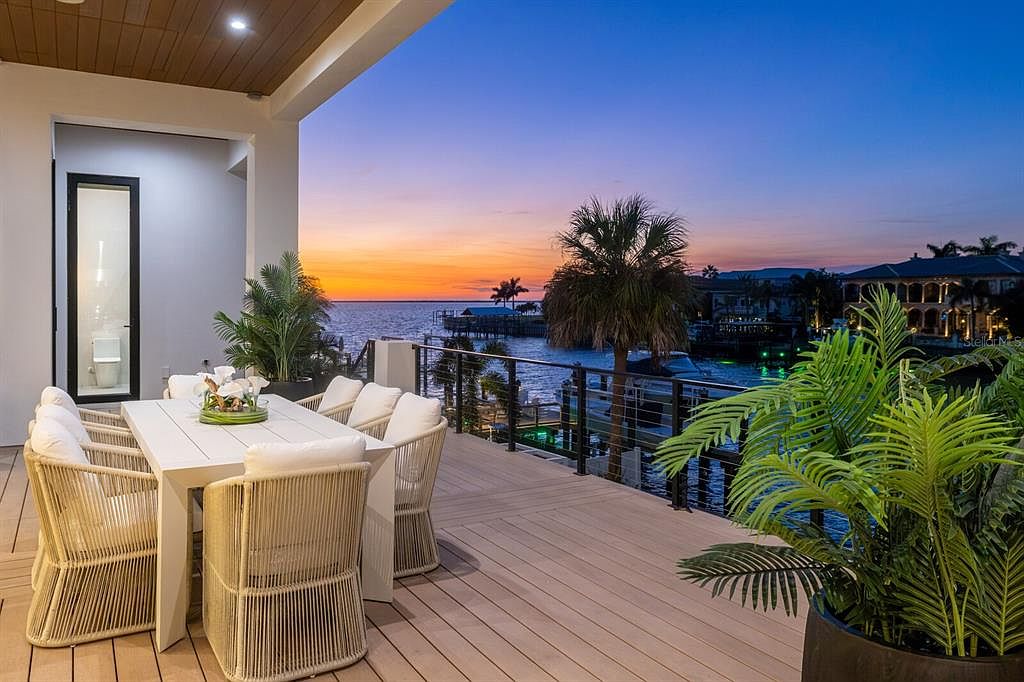 This image showcases a luxurious outdoor patio or deck area, furnished with a white dining table and woven chairs with cushions. The space overlooks a waterfront view at sunset, with palm trees and other tropical plants adding to the ambiance. The overall impression is one of relaxation and upscale outdoor living.
