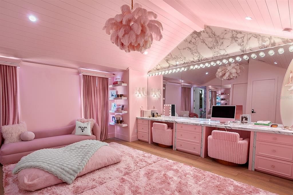 This is a pink themed bedroom featuring a plush pink rug, a pink sofa with a Prada pillow, and a large vanity area with a marble countertop and lighted mirror. The room is decorated in various shades of pink, creating a whimsical and luxurious atmosphere. The perspective is a wide angle shot to capture the entire room.