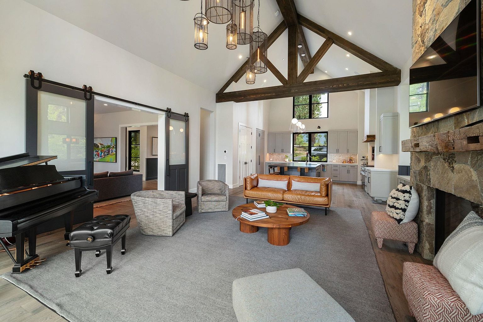 This is a spacious living room featuring a high vaulted ceiling with exposed wooden beams and a stone fireplace with a mounted TV. The room is furnished with a leather sofa, upholstered chairs, a round wooden coffee table, and a grand piano. Sliding barn doors lead to another room, and the open floor plan seamlessly connects to the kitchen, creating a bright and inviting atmosphere.