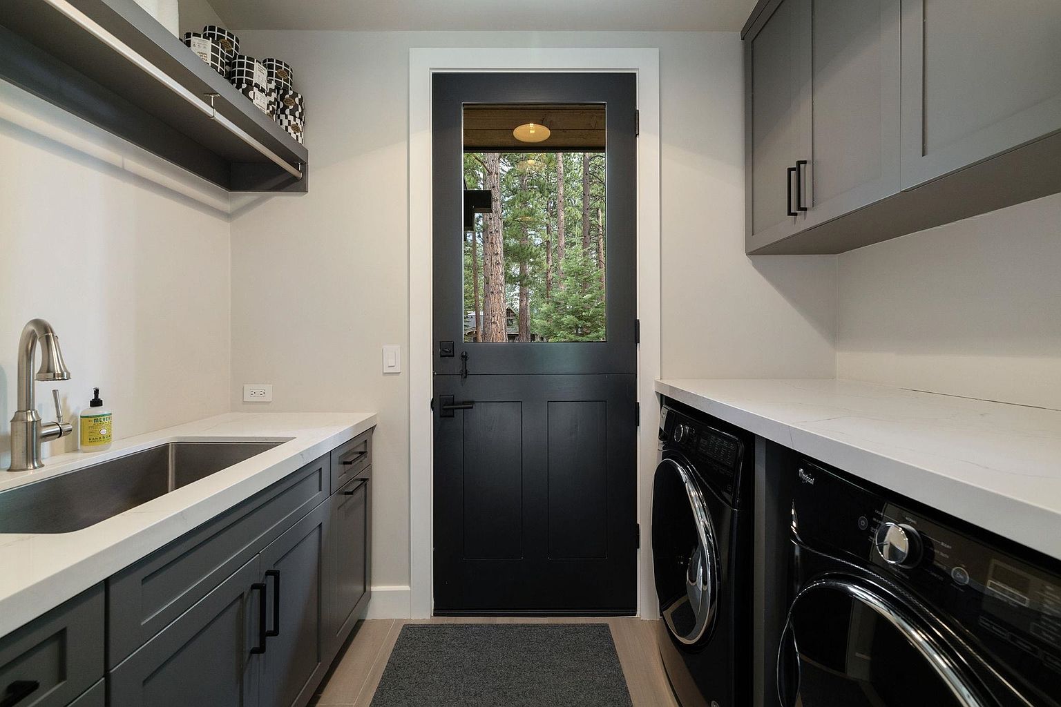 This is a well-organized laundry room featuring a dark gray color scheme with white countertops. A stainless steel sink and faucet are on the left, while a black washer and dryer are on the right. A black Dutch door with a window provides natural light and a view of the outdoors, and gray cabinets above the appliances offer ample storage space.