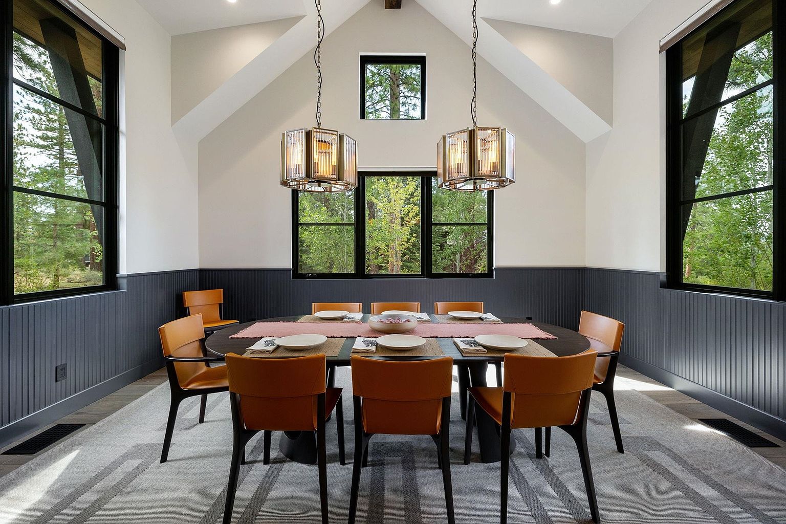 This is an interior shot of a dining room featuring a long, oval-shaped table set for a meal, surrounded by tan leather chairs with black legs. The room has a modern farmhouse aesthetic with black-framed windows offering views of the surrounding greenery, and two pendant lights hang above the table, adding a touch of elegance. The walls are two-toned, with a dark gray wainscoting and light beige above, creating a sophisticated and inviting atmosphere.