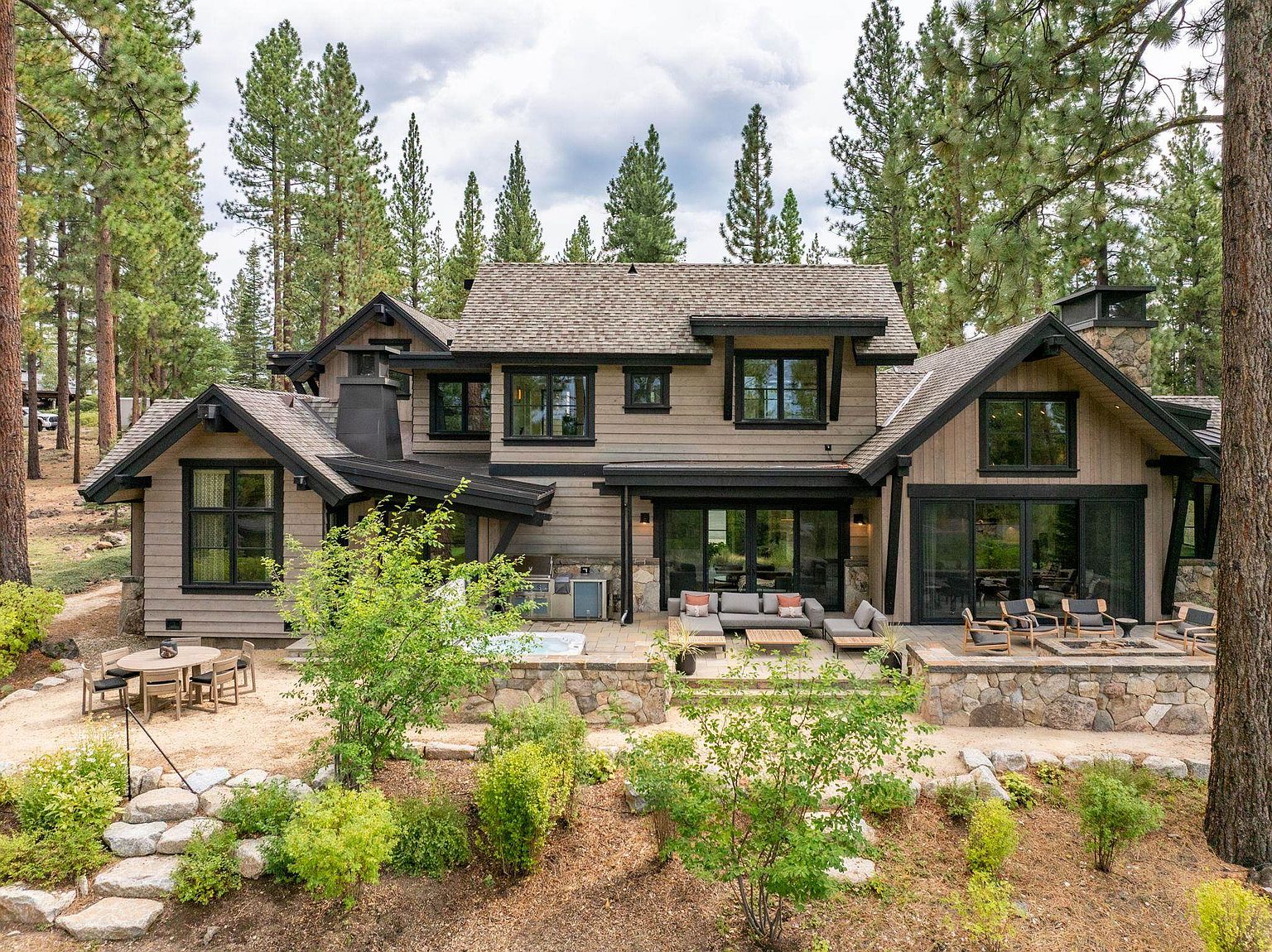 This image showcases the rear exterior of a luxurious home, featuring a multi-level stone patio with outdoor seating and a fire pit. The house has a combination of wood and stone accents, with large windows and sliding glass doors that provide seamless indoor-outdoor living. Mature trees surround the property, creating a sense of privacy and tranquility.