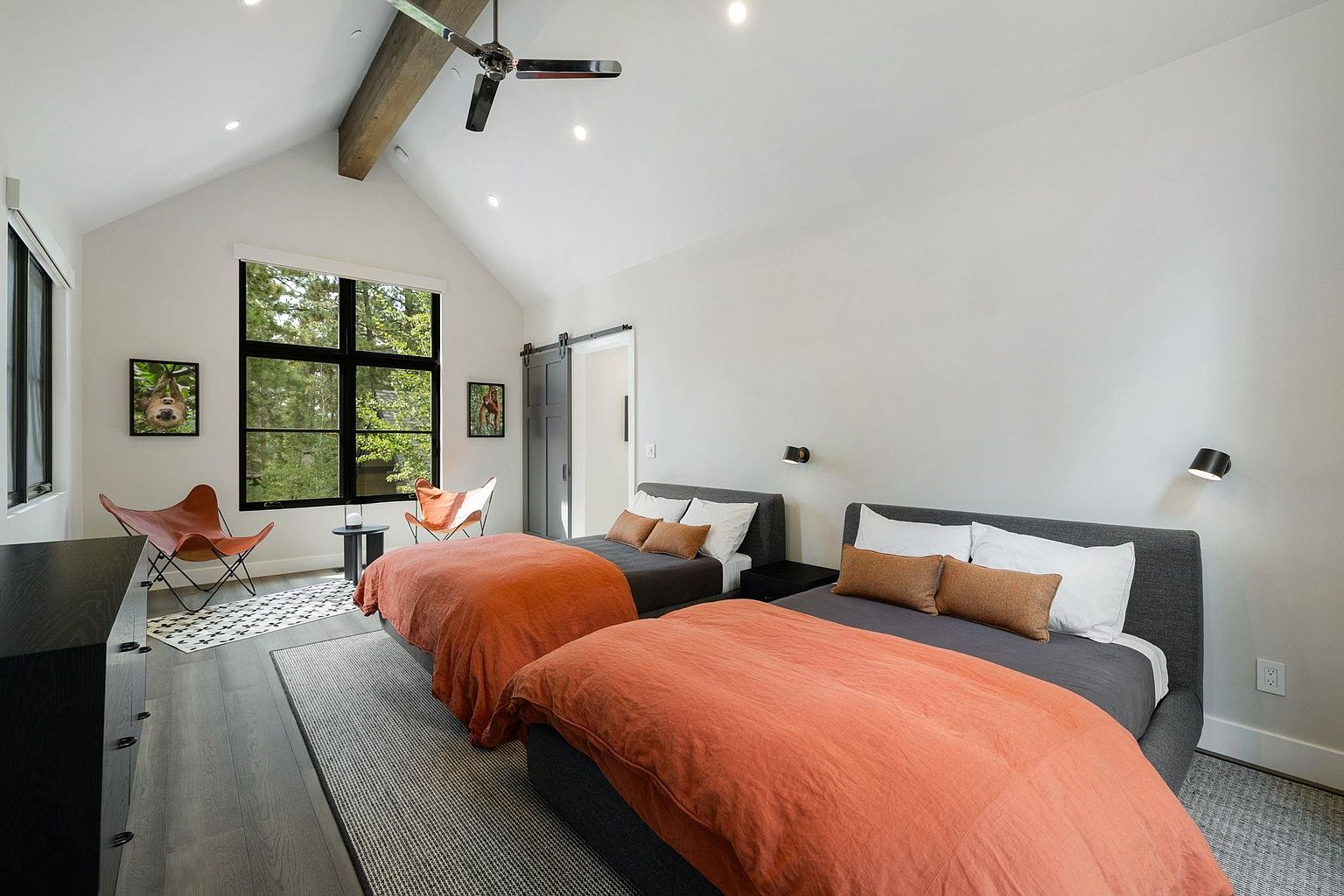 This is a bright and airy guest bedroom featuring two beds with gray frames and orange bedding. The room has a modern aesthetic with white walls, a vaulted ceiling with a wooden beam, and a large window offering a view of the surrounding greenery. The room is decorated with butterfly chairs and artwork, creating a cozy and inviting atmosphere.
