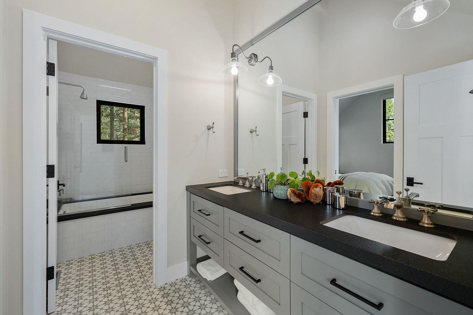 This is a well-lit bathroom featuring a double vanity with a dark countertop and gray cabinetry. The floor is tiled with a geometric pattern, and there's a large mirror above the vanity. Through the doorways, we can see a shower/tub combo and a bedroom, adding depth and context to the space.