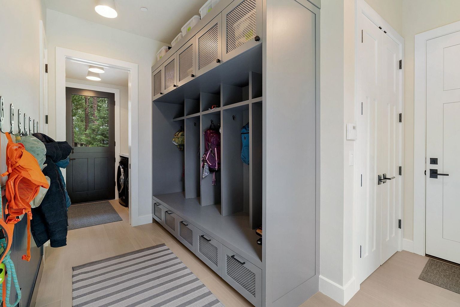 This interior shot showcases a well-organized mudroom area with a custom gray built-in storage unit featuring lockers, drawers, and overhead compartments. The space is bright and clean, with light wood flooring and a striped rug adding visual interest. A glimpse of the outdoors is visible through a nearby door, enhancing the sense of connection to nature.