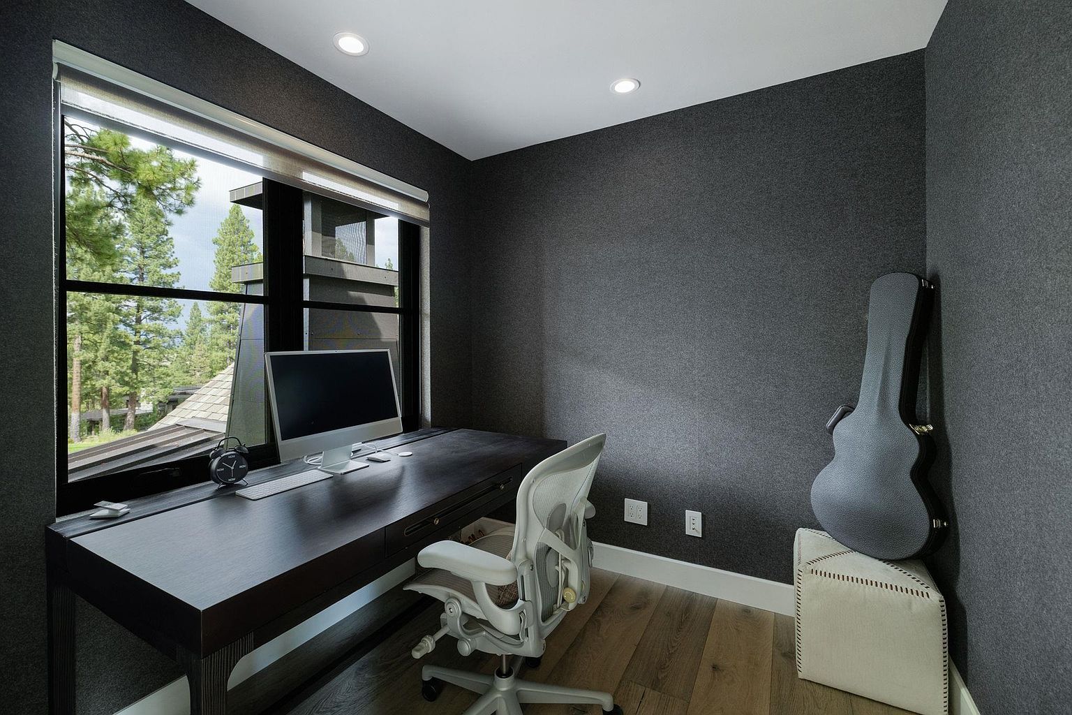 This is an interior shot of a modern office space. The room features a dark wood desk with a computer setup, a comfortable ergonomic chair, and a guitar case leaning against the wall on top of a white ottoman. A large window provides natural light and a view of the outdoors, creating a serene and productive atmosphere.