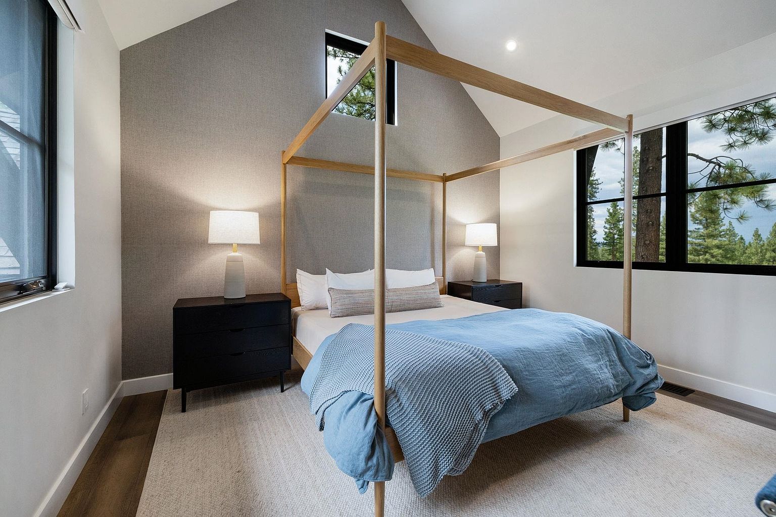 This is a bright and airy primary bedroom featuring a four-poster bed with a light blue duvet and a textured throw. The room has a neutral color palette with a textured gray accent wall and white walls, complemented by dark wood nightstands and matching lamps. Large windows provide natural light and views of the outdoors, enhancing the serene and inviting atmosphere.