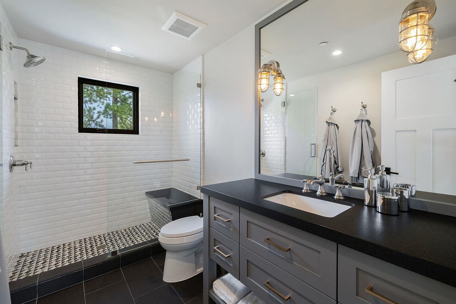 This is a well-lit bathroom featuring a modern design with a gray vanity topped with a black countertop and a white undermount sink. The shower area is enclosed with glass and features white subway tiles and a black and white patterned floor. A black-framed window provides natural light, and the overall impression is clean and stylish.