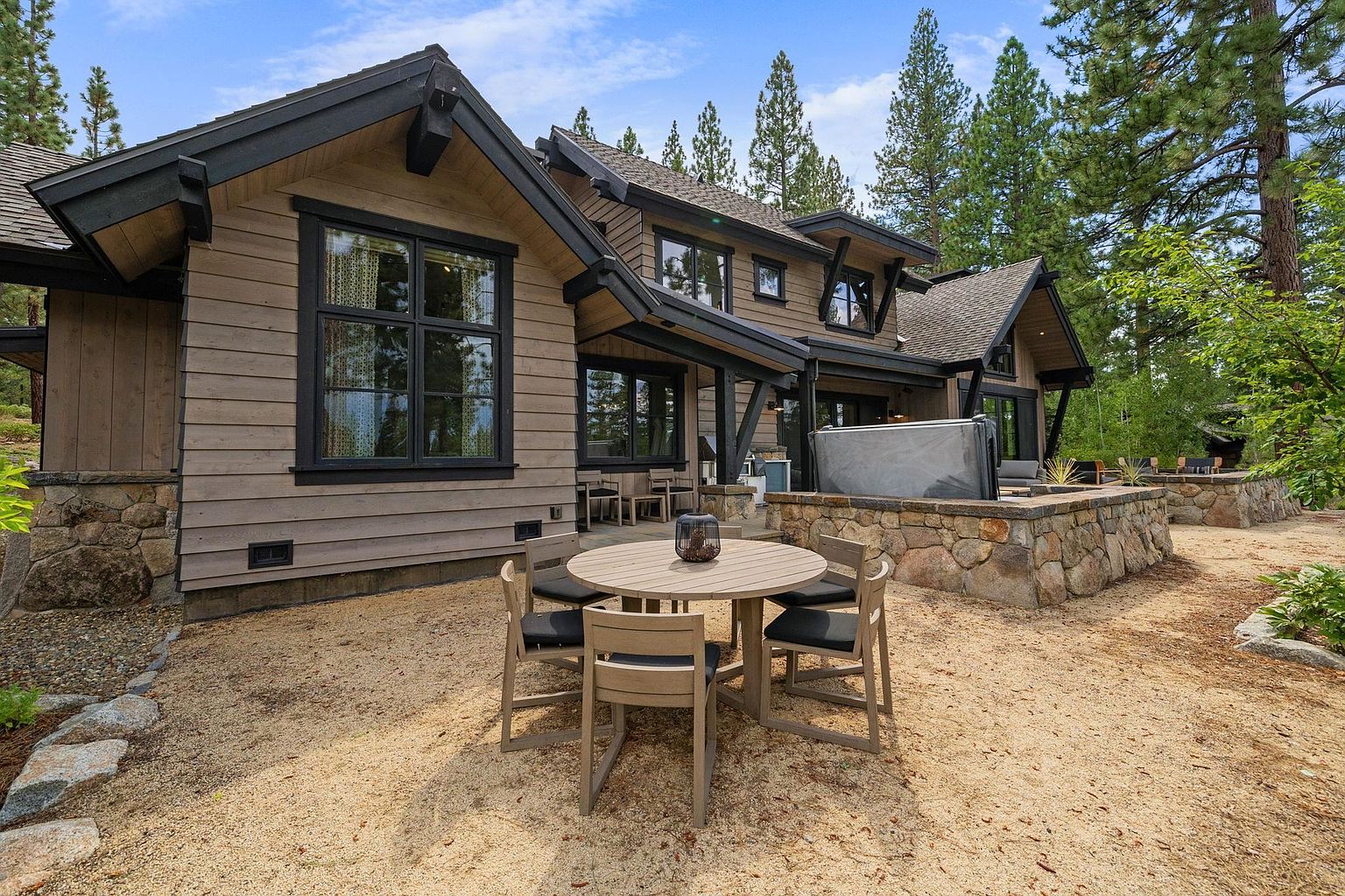 This image showcases a charming outdoor patio area of a home, featuring a round wooden table with four chairs, set on a gravel surface. The patio is adjacent to a stone-clad structure, possibly housing a hot tub, and the backdrop includes a well-maintained wooden house with black-framed windows and lush greenery. The scene evokes a sense of relaxation and outdoor living, perfect for entertaining or enjoying quiet moments.