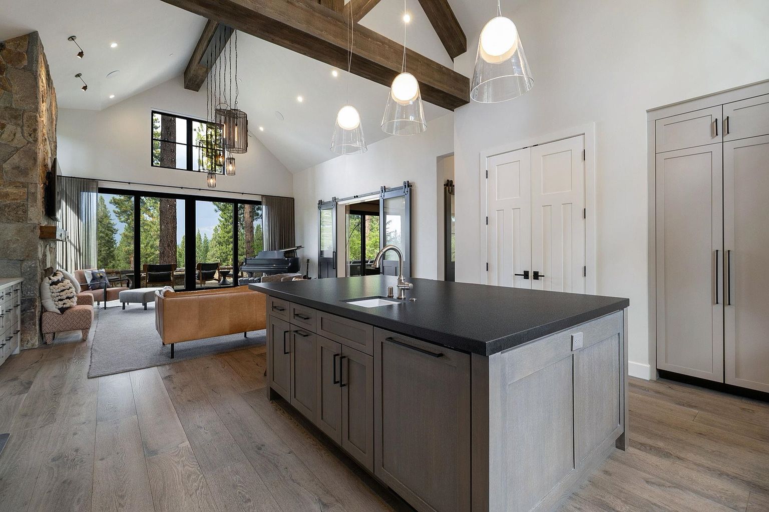 This interior shot showcases a modern kitchen and living space with an open floor plan. The kitchen features a large island with a dark countertop and gray cabinetry, complemented by pendant lighting. The living area includes a stone fireplace, comfortable seating, and large windows offering a view of the outdoors, creating a bright and inviting atmosphere.