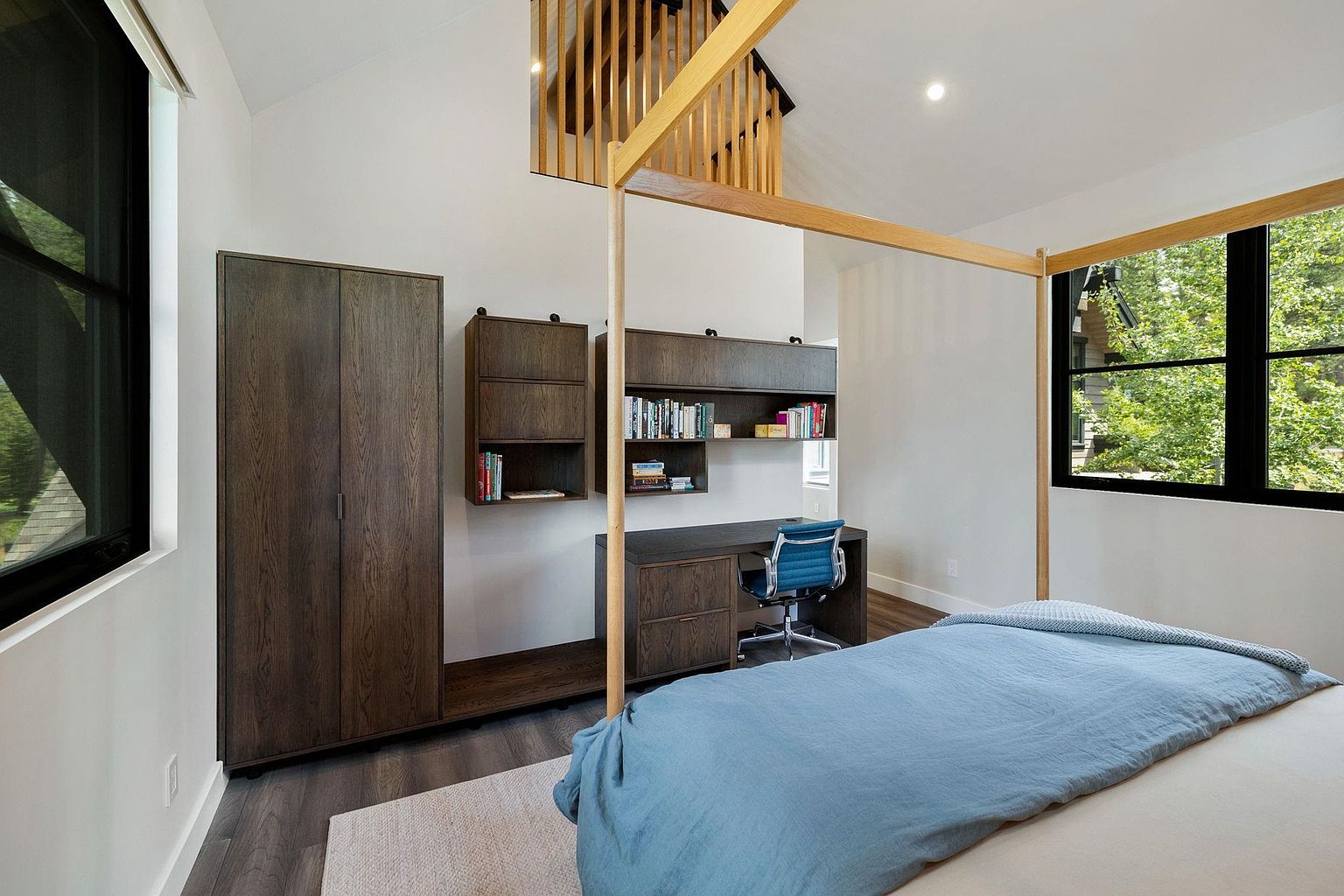 This is a bright and modern primary bedroom featuring a unique four-poster bed frame, a built-in desk and shelving unit, and a spacious wardrobe. The room is well-lit with natural light from a large window, and the color palette is neutral with pops of blue. The overall impression is clean, contemporary, and inviting.