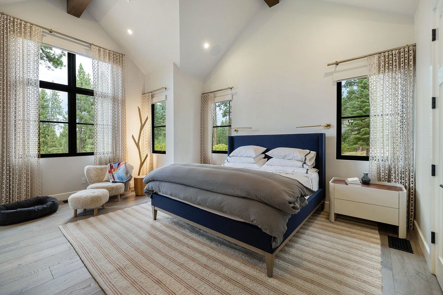 This is a primary bedroom featuring a blue upholstered bed with white linens and a gray duvet. The room has a high, vaulted ceiling and is illuminated by natural light streaming through large windows framed by patterned curtains. A striped rug covers the light wood floor, and a small seating area with a chair and ottoman is visible near one of the windows, creating a cozy and inviting atmosphere.