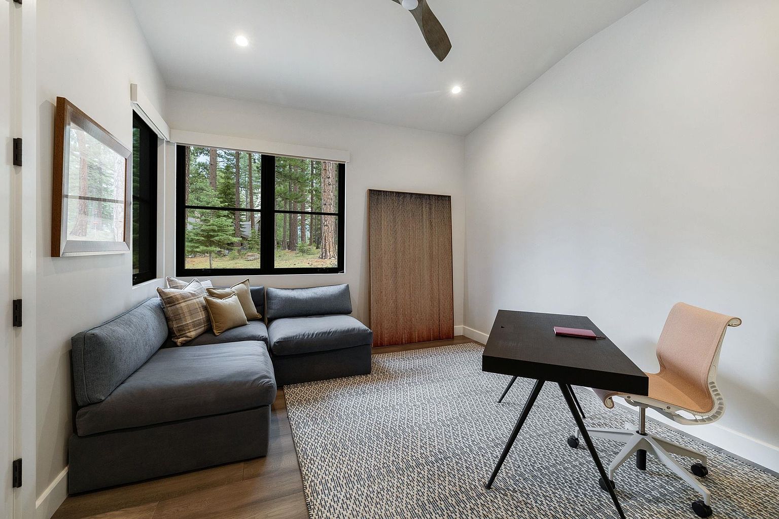 This is an interior shot of a modern office or study. The room features a gray sectional sofa with throw pillows, a black desk with a chair, and a large window offering a view of a forest. The walls are white, and the flooring is a combination of wood and a patterned rug, creating a clean and inviting workspace.