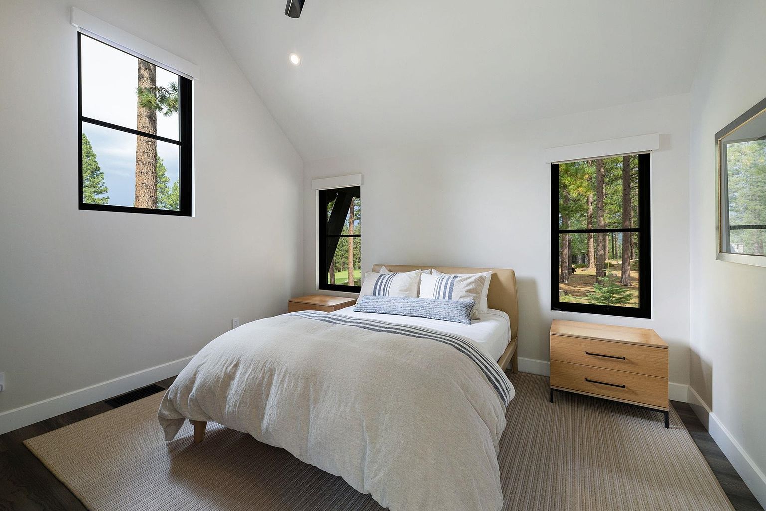 This is a bright and airy bedroom featuring a neutral color palette. The room has three windows with black frames, offering views of the surrounding trees. A bed with a light-colored duvet and striped pillows is the focal point, complemented by wooden nightstands on either side. The room is carpeted and has a minimalist, modern aesthetic.