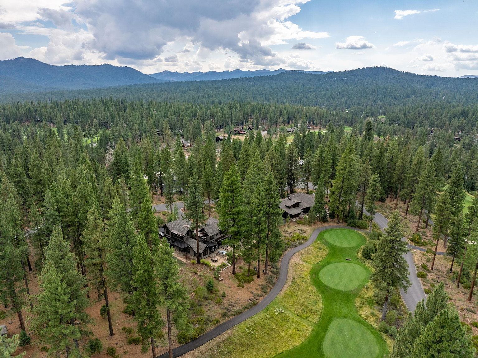 This aerial shot showcases a luxurious property nestled among tall pine trees, adjacent to a well-manicured golf course. The home features a dark roof and blends seamlessly with the natural surroundings, offering a sense of privacy and exclusivity. The landscape includes lush greenery and a winding road leading to the property, with mountains visible in the distance.