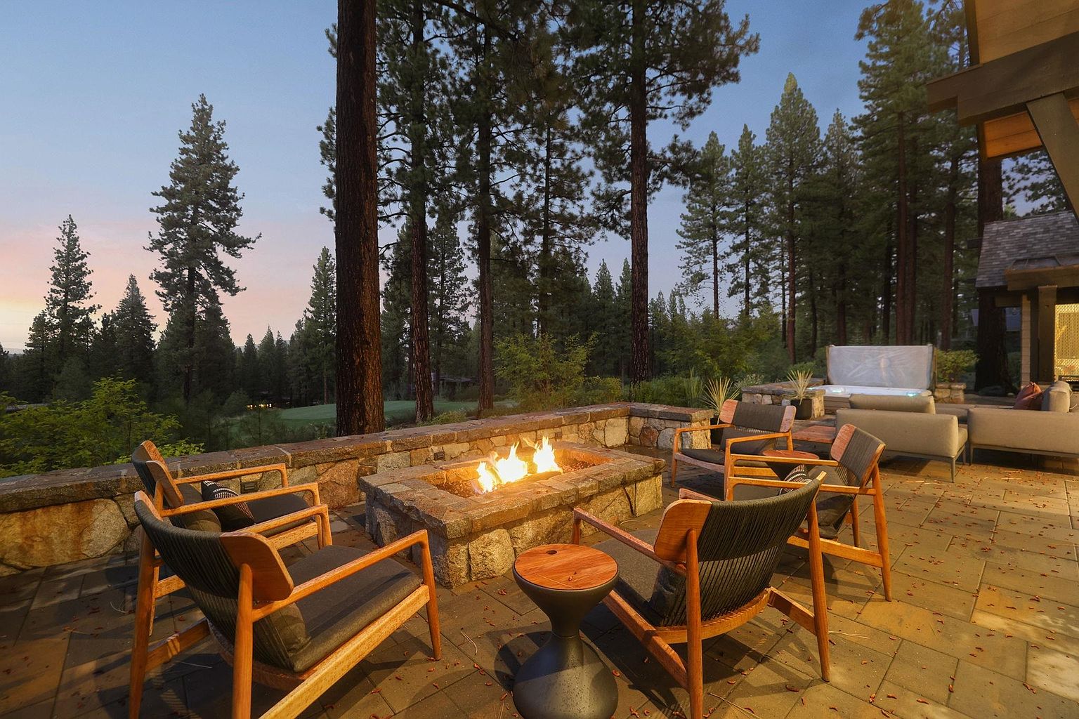 This image showcases a beautifully designed outdoor patio area, featuring a stone fire pit surrounded by comfortable wooden chairs with dark cushions. The patio is paved with stone tiles and is adjacent to a well-maintained lawn and tall trees, creating a serene and inviting atmosphere. In the background, there is a glimpse of a hot tub and outdoor seating, suggesting a luxurious and relaxing outdoor living space.