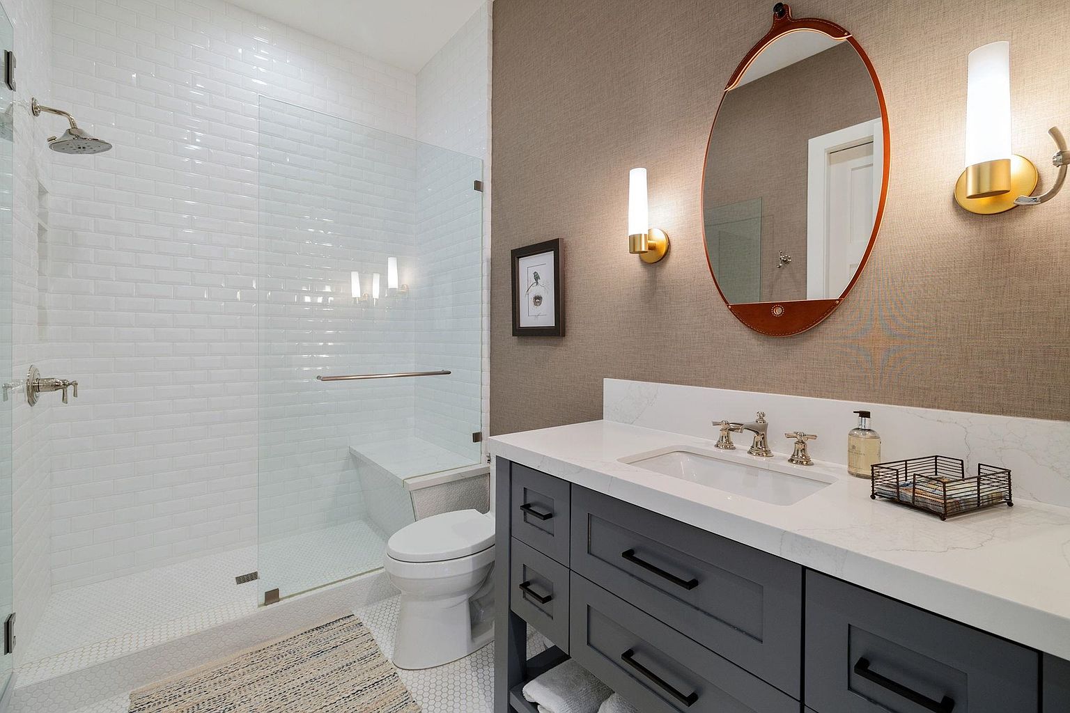 This is a well-lit bathroom featuring a walk-in shower with white subway tile and a glass enclosure. The vanity has a gray cabinet with a white countertop, a rectangular sink, and modern fixtures. An oval mirror with leather trim hangs above the vanity, flanked by sconce lighting, and the walls are covered in a textured wallpaper.