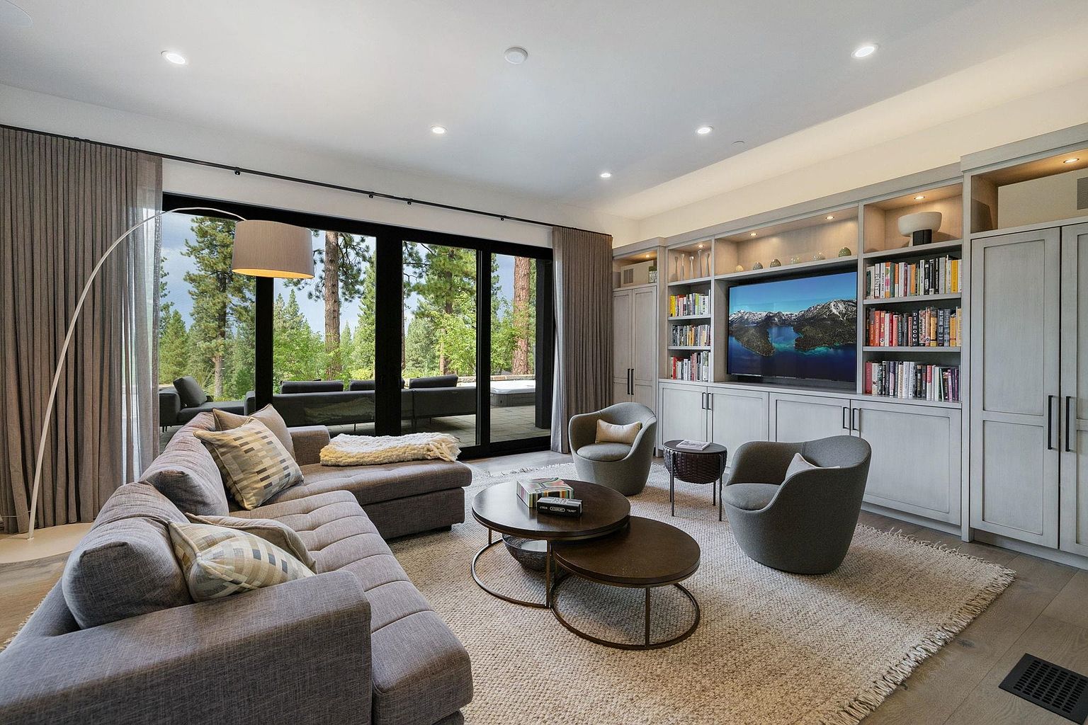 This is a well-lit living room featuring a large gray sectional sofa with decorative pillows, two round coffee tables, and two gray armchairs. A built-in shelving unit with a television is against one wall, and a large sliding glass door offers a view of the outdoor scenery. The room is decorated in neutral tones with a focus on comfort and modern design.