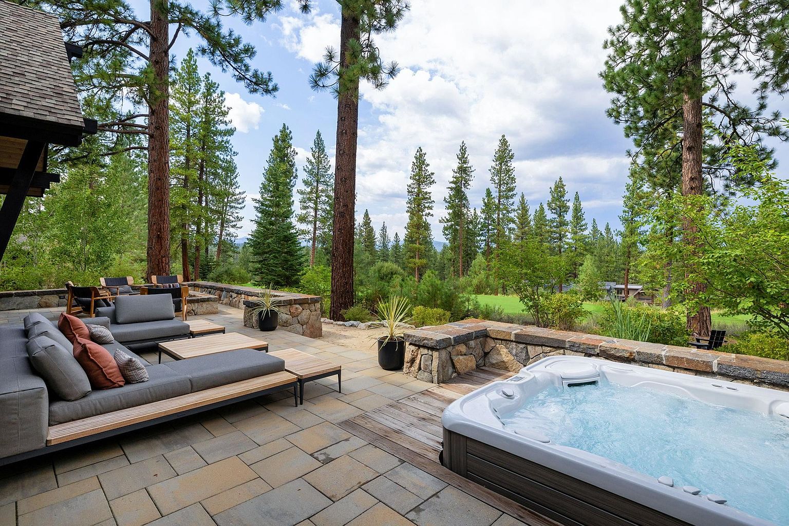 This image showcases a luxurious outdoor living space featuring a spacious patio with stone pavers, comfortable lounge seating, and a hot tub. The patio is surrounded by mature trees and a stone retaining wall, creating a private and serene atmosphere. The overall impression is one of relaxation and upscale outdoor living.