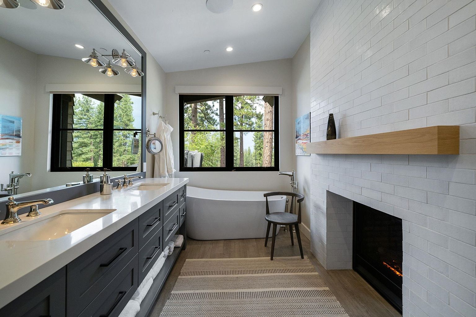 This is a luxurious primary bathroom featuring a double vanity with gray cabinetry and white countertops, a freestanding bathtub positioned under a window with a view of trees, and a fireplace with a white brick surround and a wooden mantel. The room is well-lit with recessed lighting and stylish fixtures, creating a spa-like atmosphere.