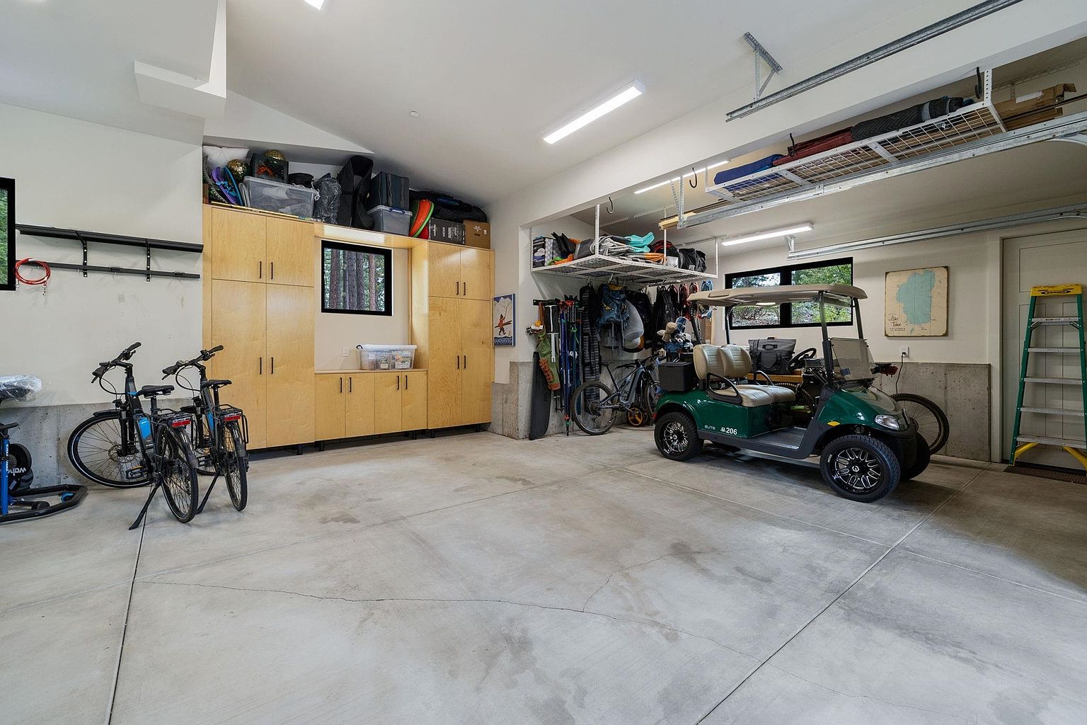 This is a well-organized garage featuring built-in wooden cabinets along one wall, providing ample storage space. A golf cart is parked in the center, and bicycles are neatly arranged to the side. Overhead storage racks maximize vertical space, and the concrete floor appears clean and well-maintained, suggesting a functional and tidy area.