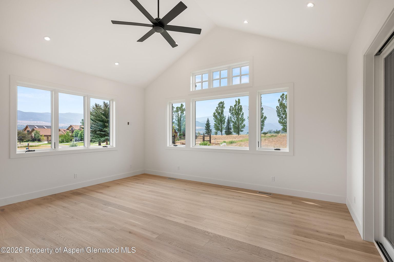 This bright and airy living room features vaulted ceilings, light wood flooring, and crisp white walls that create a clean, modern aesthetic. Large windows on two walls provide abundant natural light and expansive views of the surrounding landscape. A dark ceiling fan serves as a central fixture, adding a touch of contrast to the minimalist, open-concept space.