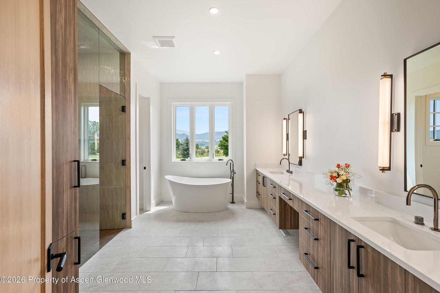 This spacious primary bathroom features a clean, modern aesthetic with a long dual-sink vanity topped with light-toned stone and accented by warm wood cabinetry. A freestanding soaking tub is positioned centrally in front of a large window that provides natural light and scenic views, while a glass-enclosed shower area is located to the left. The room is finished with neutral large-format floor tiles and minimalist wall sconces, creating a bright and serene atmosphere.