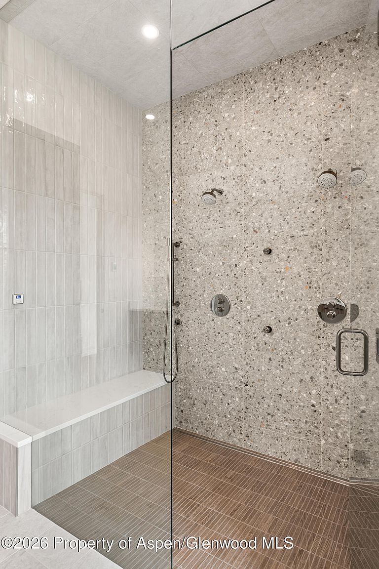 This modern walk-in shower features a striking terrazzo-style wall finish contrasted by vertical light-toned tiles on the adjacent wall. A built-in bench provides seating, while a glass partition separates the shower area from the rest of the room, which is finished with linear brown floor tiles. The space is equipped with multiple shower heads and controls, creating a luxurious and functional spa-like atmosphere.