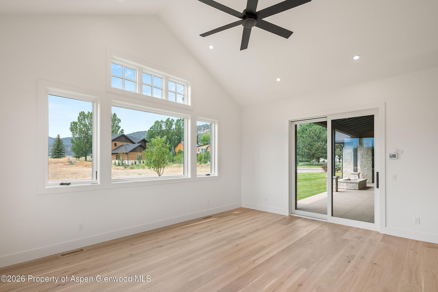 This bright, minimalist living room features light wood flooring and crisp white walls, creating an airy and open atmosphere. Large windows and a sliding glass door provide abundant natural light and views of the surrounding landscape and patio. A modern ceiling fan is centered in the vaulted ceiling, adding a functional and sleek design element to the space.