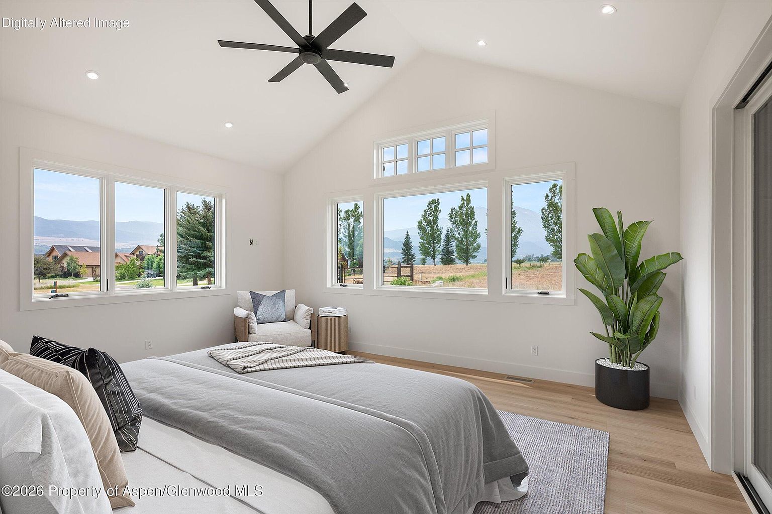 This bright and airy primary bedroom features a vaulted ceiling with a modern ceiling fan and light-toned wood flooring. Large windows provide expansive views of the surrounding landscape, while a cozy armchair and a potted plant add comfort to the minimalist space. The room is anchored by a bed with neutral bedding, creating a serene and inviting atmosphere.
