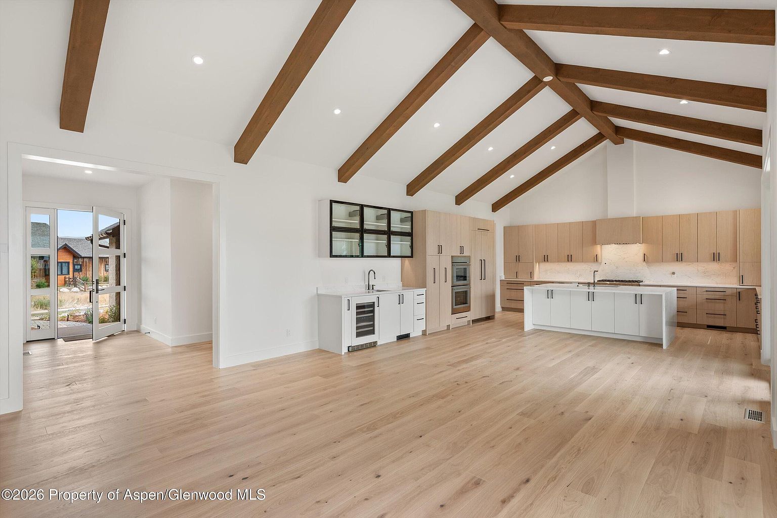 This spacious, open-concept kitchen features a vaulted ceiling with exposed wooden beams and light-toned wood flooring throughout. The area includes a large central island with a white base, light-colored cabinetry, and a dedicated wet bar section with a wine refrigerator. A wide doorway leads to an exterior entryway, creating a bright and airy atmosphere with a modern, minimalist aesthetic.