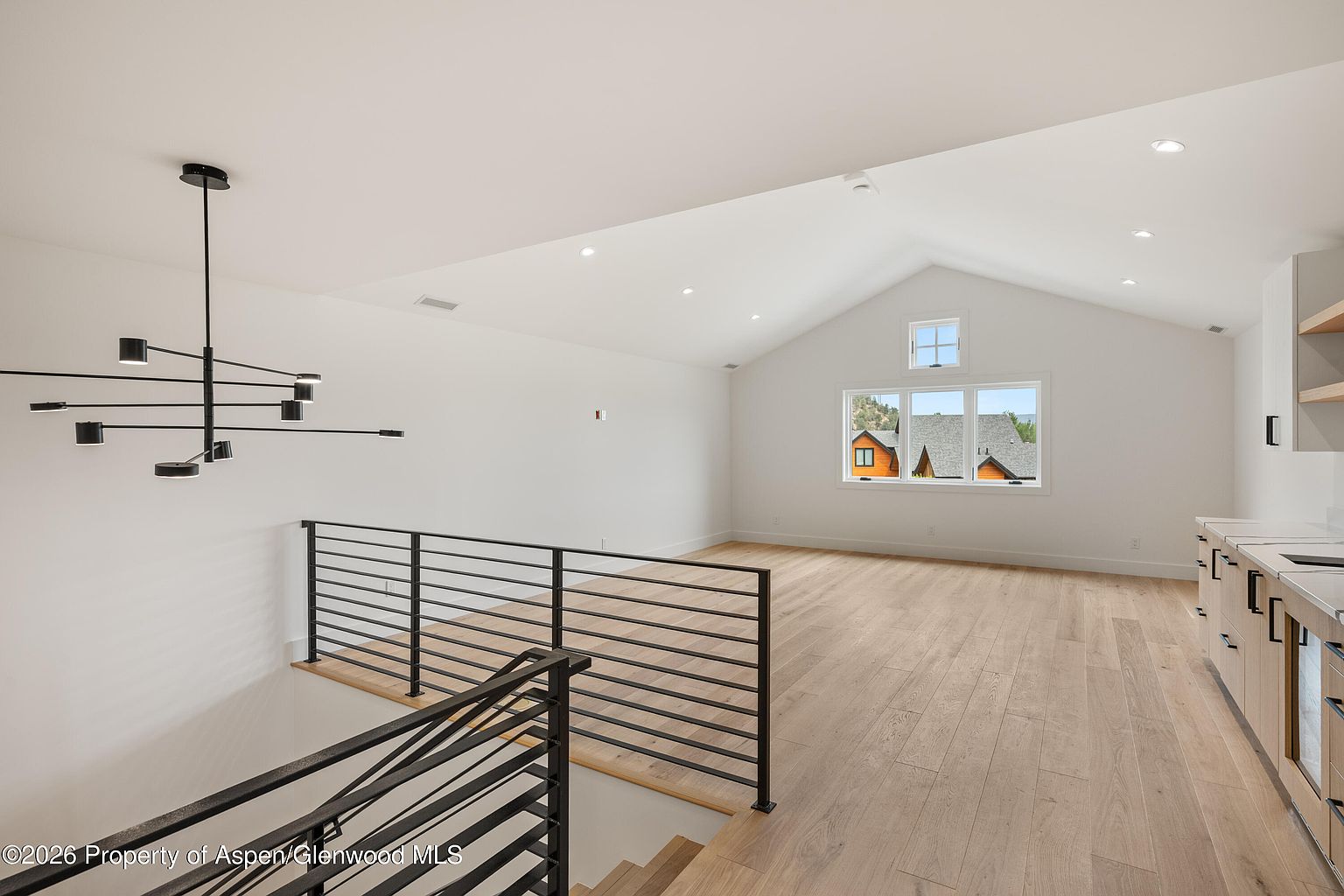 This bright, open-concept living area features vaulted ceilings and light wood flooring, creating an airy and modern atmosphere. A striking black multi-arm chandelier hangs above the staircase, which is enclosed by a sleek black metal railing. Large windows provide natural light and a view of the surrounding landscape, while a partial kitchen area is visible on the right side of the frame.