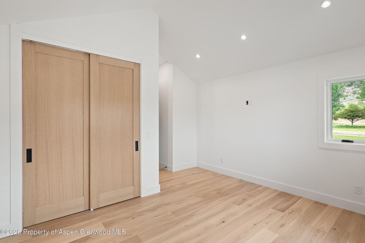 This bright, minimalist bedroom features light wood flooring and crisp white walls, creating an airy and clean aesthetic. A set of sliding light wood doors with black hardware provides access to a closet, while a single window offers a view of the surrounding landscape. The room is illuminated by recessed ceiling lights, emphasizing the simple, modern design.
