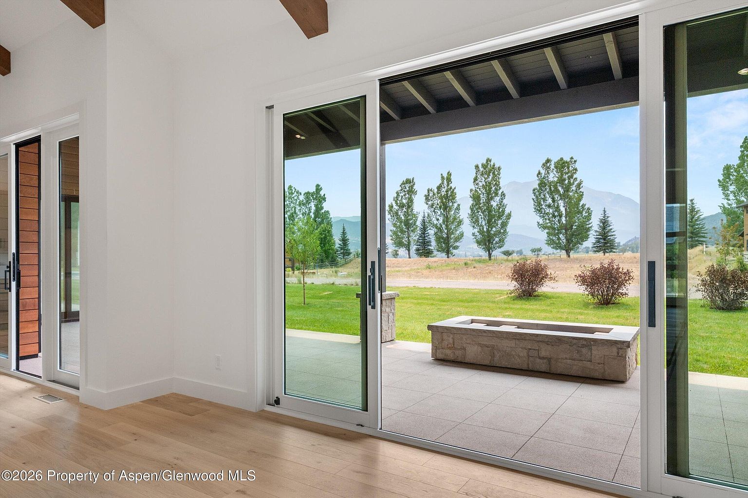 This interior view captures a bright living space transitioning seamlessly to an outdoor patio through large sliding glass doors. The room features light wood flooring and white walls, while the exterior showcases a stone fire pit on a paved patio overlooking a lush green lawn and distant mountains. The perspective emphasizes the indoor-outdoor connection, highlighting the expansive views and natural light.