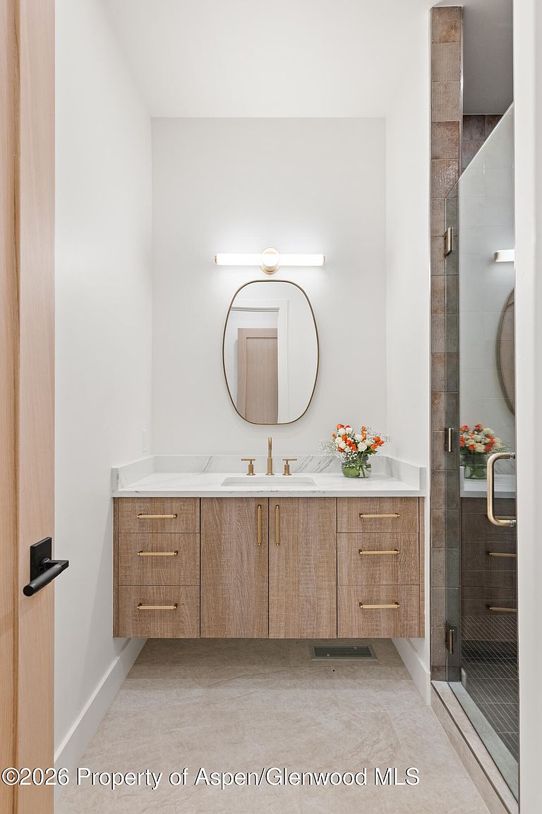 This modern bathroom features a floating vanity with light wood cabinetry and gold-toned hardware, topped with a white stone countertop. An oval mirror is centered above the vanity, illuminated by a sleek, horizontal light fixture, while a glass-enclosed shower with textured tile accents sits to the right. The space is finished with neutral-toned flooring and white walls, creating a clean and contemporary aesthetic.