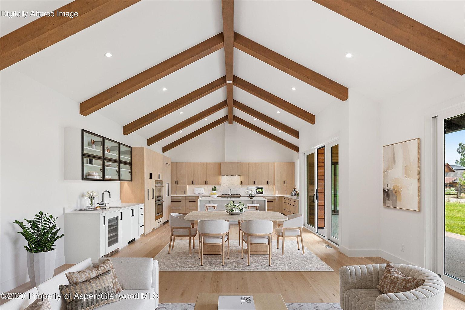 This open-concept kitchen and dining area features a vaulted ceiling with exposed wood beams and light wood cabinetry. A central island with bar seating sits in front of a dining table, while a wet bar with glass-front cabinets is positioned to the left. Large glass doors provide a view of the outdoor space, creating a bright and airy atmosphere with a modern, minimalist aesthetic.