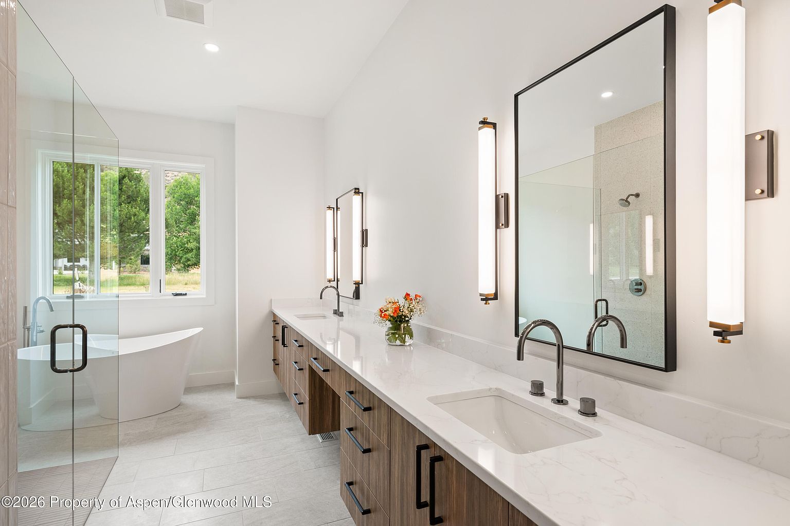 This bright and modern primary bathroom features a long dual-sink vanity with wood-toned cabinetry and a light-colored stone countertop. A large, rectangular mirror hangs above the vanity, flanked by vertical wall-mounted light fixtures, while a freestanding soaking tub sits near a window in the background. The space is finished with light-colored tile flooring and a glass-enclosed shower, creating a clean and airy atmosphere.