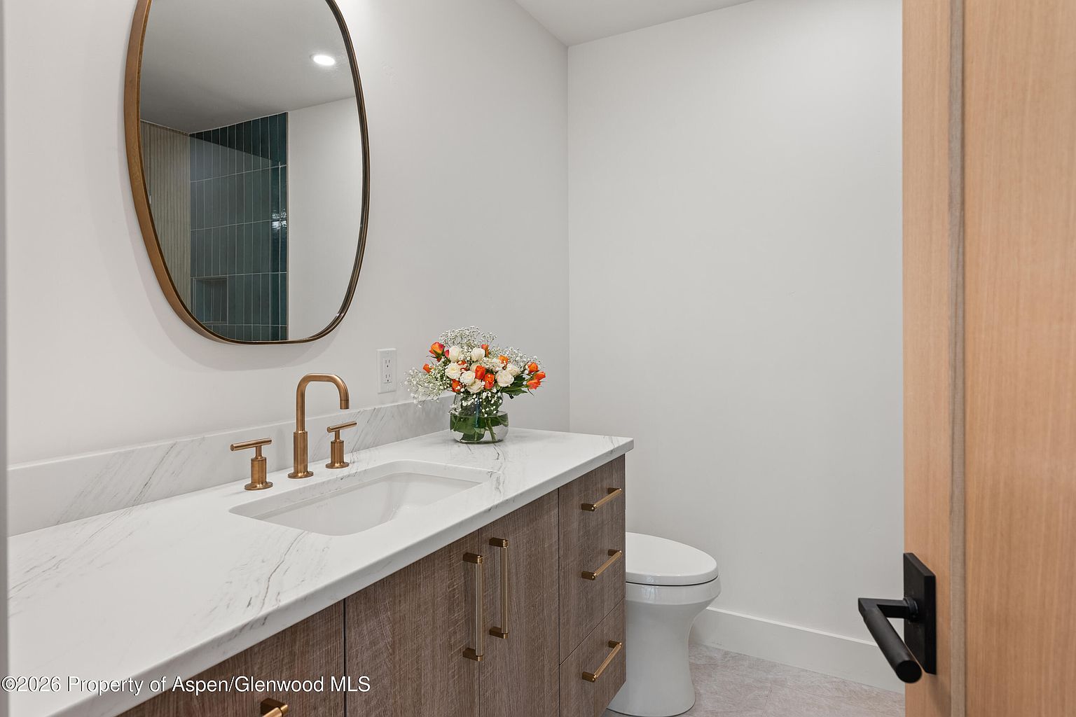 This modern powder room features a sleek vanity with a stone countertop and a gold-toned faucet. An oval mirror with a dark frame hangs above the sink, while a vase of fresh flowers adds a touch of color to the neutral space. The room is finished with textured wood-toned cabinetry and a white toilet, creating a clean and sophisticated aesthetic.