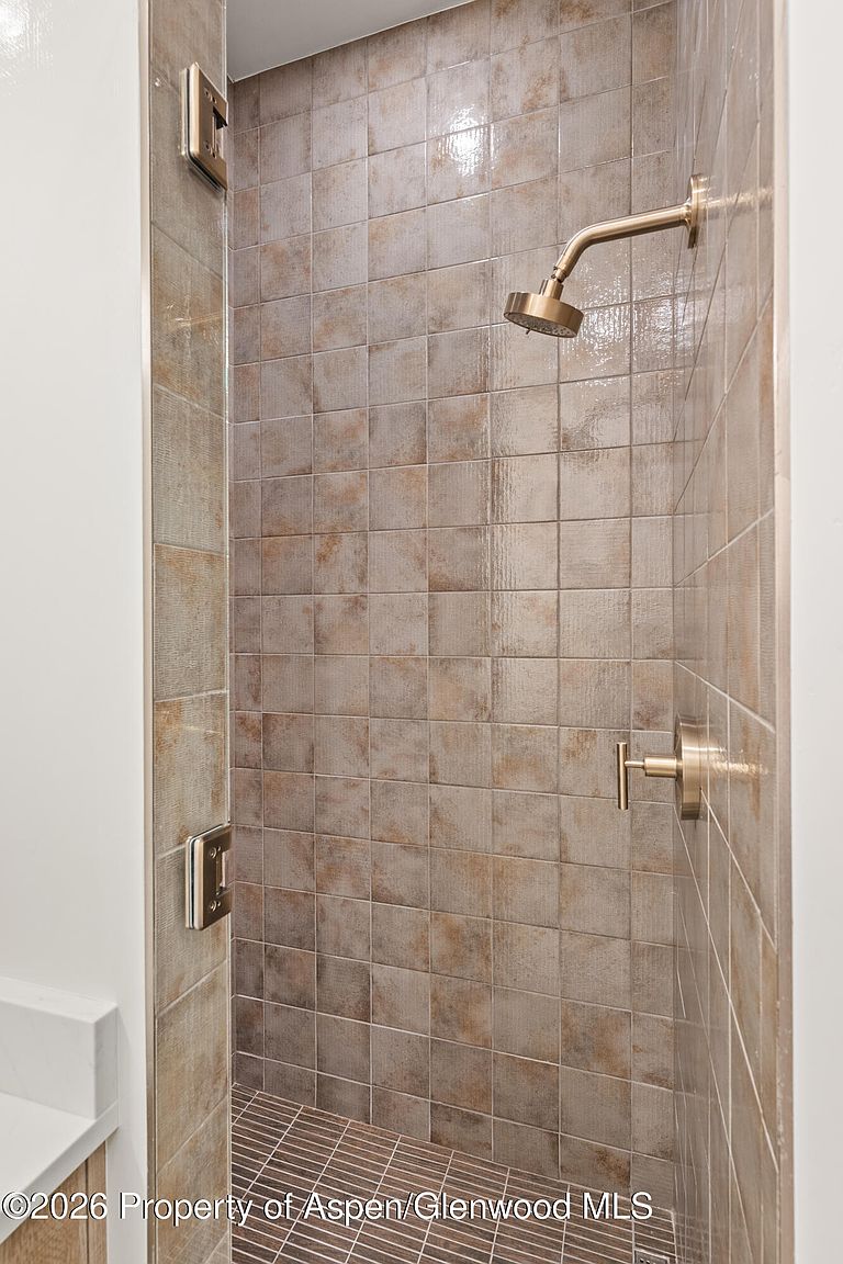 This shower stall features textured, earth-toned square tiles on the walls and a contrasting dark, linear-patterned tile on the floor. It is equipped with a modern, gold-toned shower head and matching handle fixtures, complemented by a glass door with gold-toned hardware. The perspective is a direct, eye-level shot looking into the shower enclosure, highlighting the clean, contemporary design.