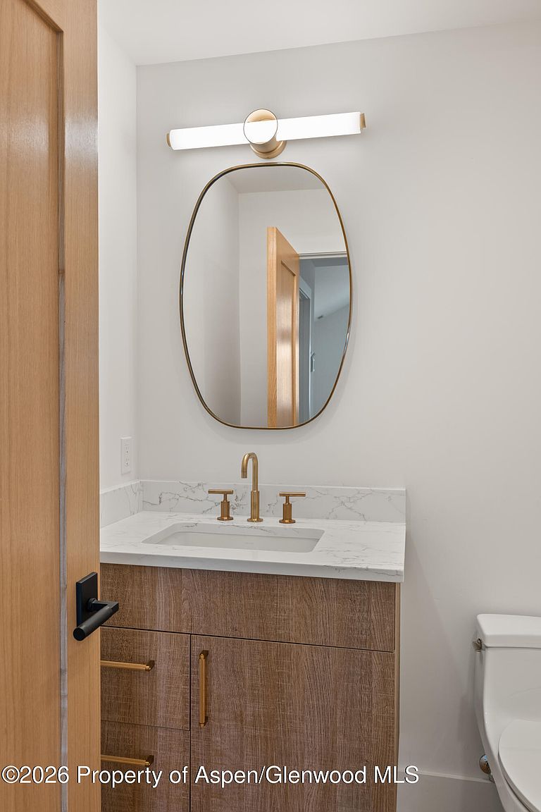 This modern powder room features a light wood vanity topped with a stone counter and a sleek, gold-toned faucet. An oval mirror with a thin gold frame hangs above the sink, illuminated by a contemporary horizontal light fixture. The space is finished with clean, neutral walls and includes a white toilet to the side, creating a bright and minimalist aesthetic.