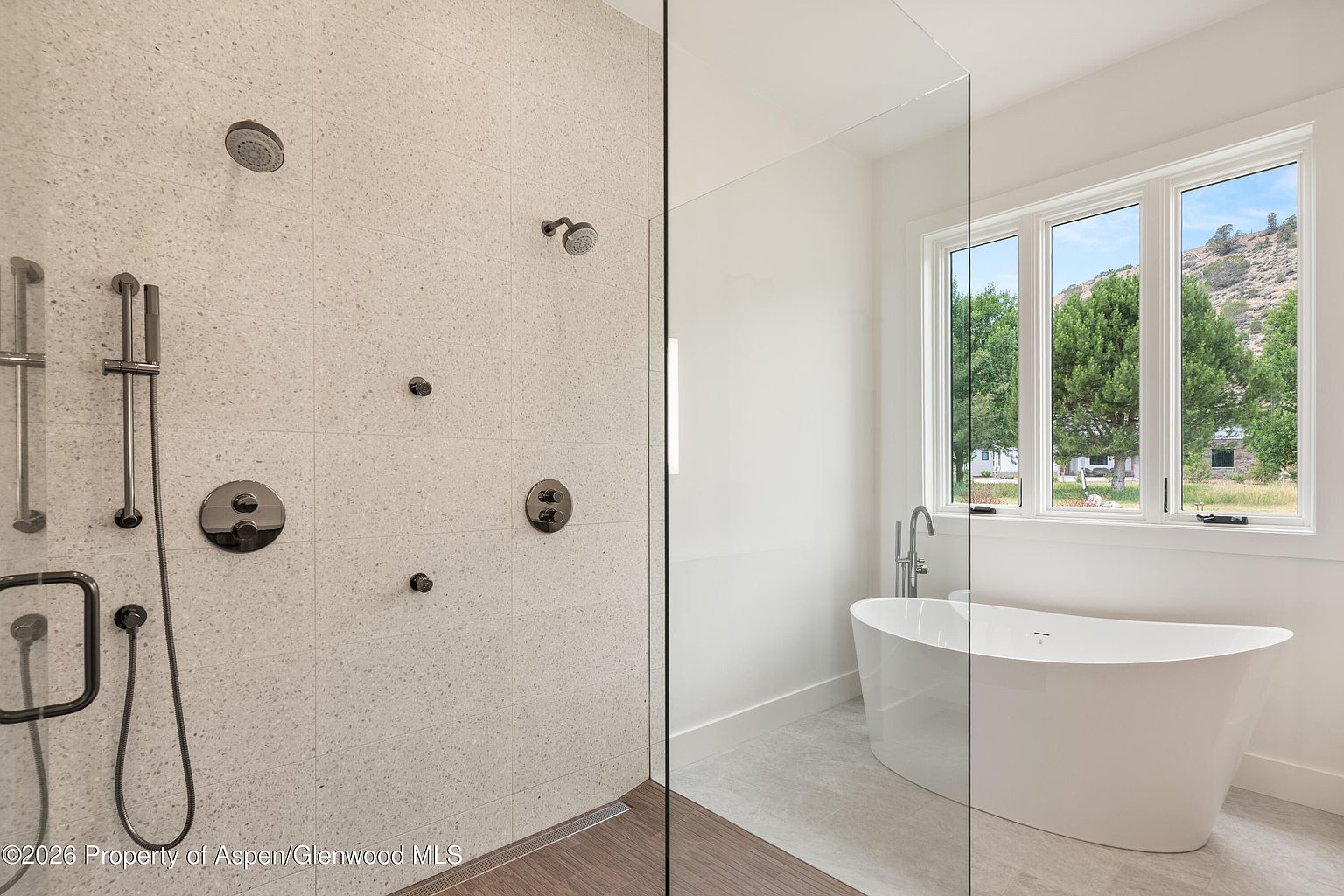 This modern primary bathroom features a spacious walk-in shower with a speckled stone-tiled wall, multiple shower heads, and a handheld sprayer. A glass partition separates the shower area from a freestanding soaking tub positioned next to large windows that offer a scenic view of the surrounding landscape. The space is bright and minimalist, utilizing a neutral color palette and clean lines to create a spa-like atmosphere.