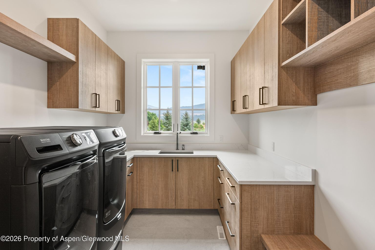 This functional laundry room features light wood cabinetry, white countertops, and a central sink positioned beneath a window that offers a scenic view of the outdoors. The space is equipped with a pair of dark-toned, front-loading laundry appliances and provides ample storage with both upper cabinets and lower drawers. The clean, neutral aesthetic creates a bright and organized workspace.