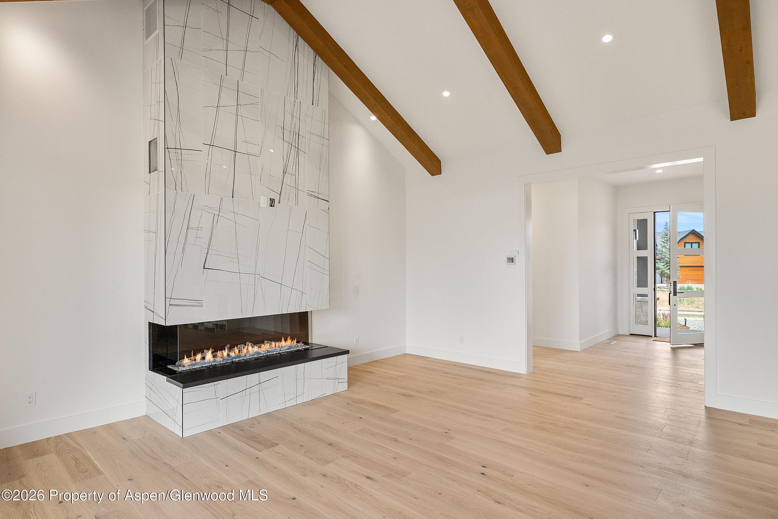 This bright living room features a vaulted ceiling accented by exposed wooden beams and a prominent fireplace with a tall, patterned stone surround. The space is finished with light-toned wood flooring and crisp white walls, creating an airy and modern atmosphere. A wide doorway in the background provides a view toward the home's entryway and exterior, emphasizing the open-concept layout.