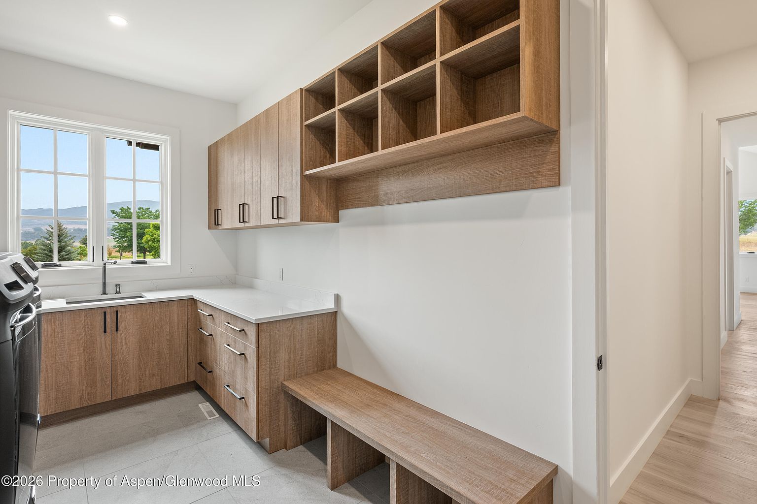 This functional laundry room features light wood-toned cabinetry with ample storage, including open cubbies and lower drawers. A white countertop provides a workspace next to a stainless steel sink, while a built-in bench offers a convenient seating area. The room is bright and airy, with a window looking out toward a scenic landscape.