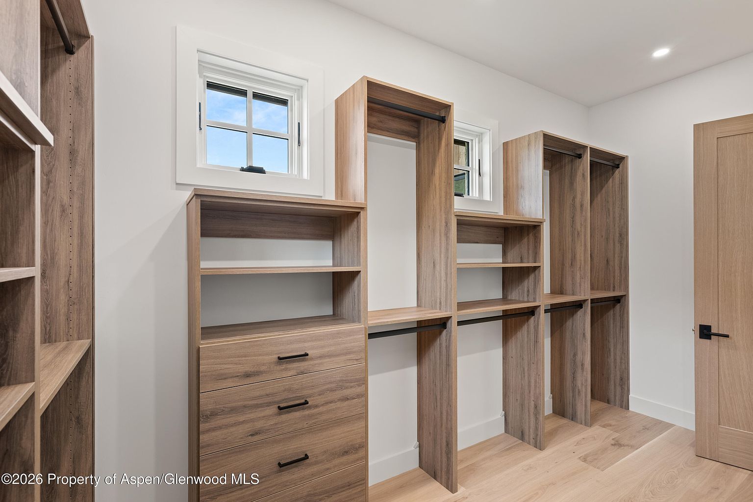 This walk-in closet features custom light wood cabinetry with a mix of open shelving, hanging rods, and a three-drawer unit. Two windows with white trim allow natural light to brighten the space, which is finished with light-toned wood flooring. The design offers a clean, organized, and modern aesthetic suitable for a primary suite.