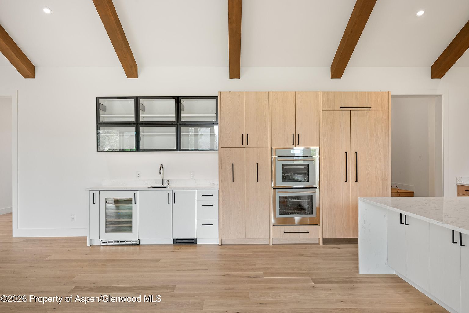 This modern kitchen features a clean, minimalist aesthetic with light wood cabinetry and white lower cabinets. The space includes a built-in double oven, a wine refrigerator, and a white stone-topped island, all set against a backdrop of exposed wooden ceiling beams. The perspective is a straight-on, eye-level shot that highlights the functional layout and the bright, airy atmosphere of the room.