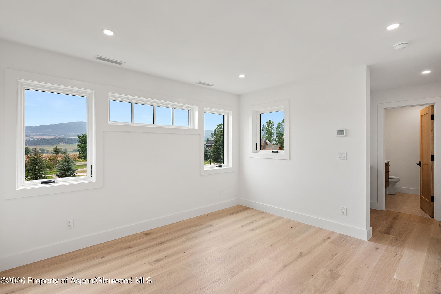 This bright, minimalist bedroom features light wood flooring and crisp white walls, creating an airy and neutral atmosphere. Large windows provide ample natural light and scenic views of the surrounding landscape, while a doorway leads to an adjacent bathroom. The clean lines and simple design offer a versatile space ready for personalization.