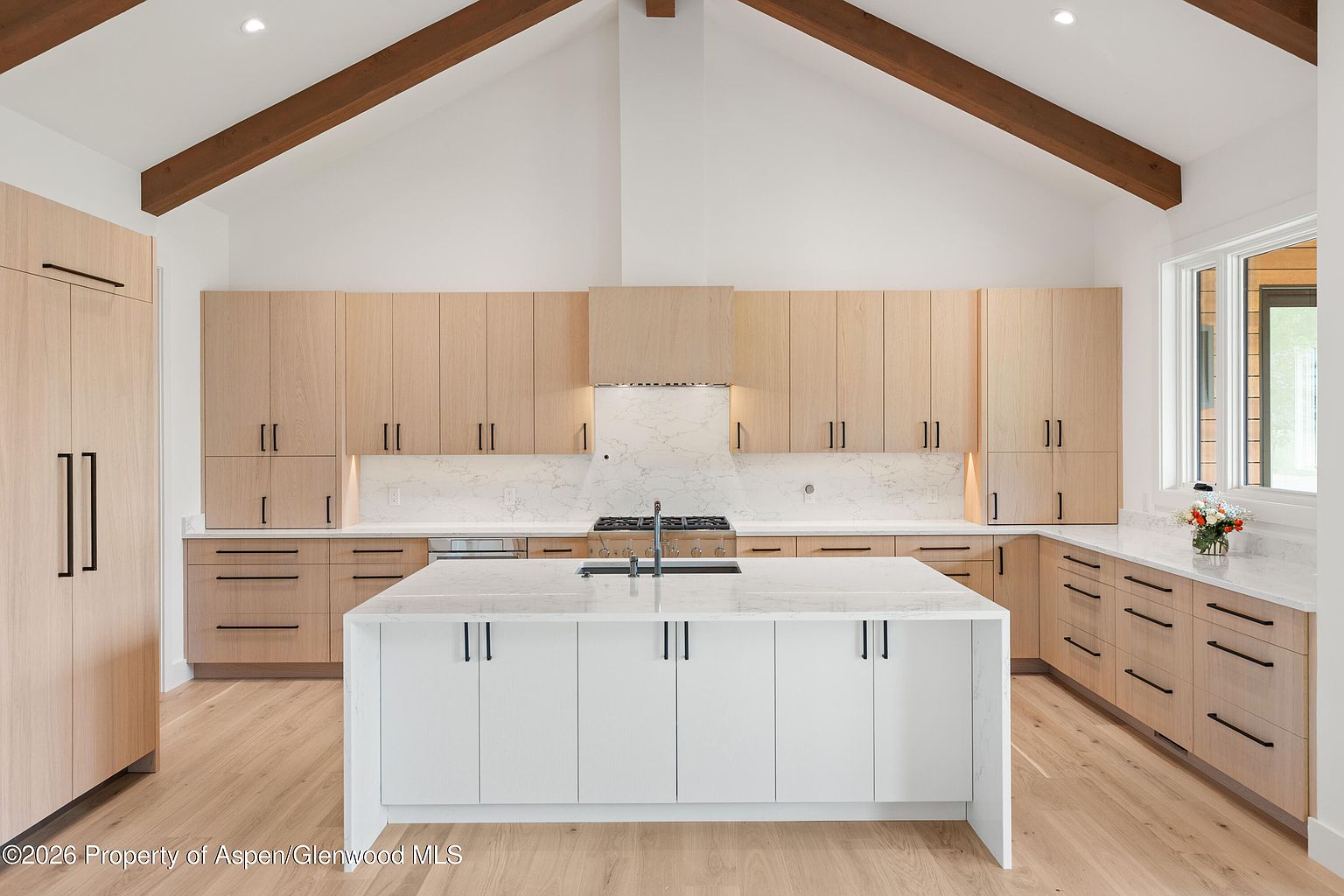 This bright, modern kitchen features a large central island with a white stone countertop and a white base, complemented by light wood cabinetry throughout. The space is defined by a vaulted ceiling with exposed wooden beams and a clean, minimalist aesthetic. A white stone backsplash behind the range adds a subtle texture, while the overall design emphasizes a spacious, airy atmosphere.