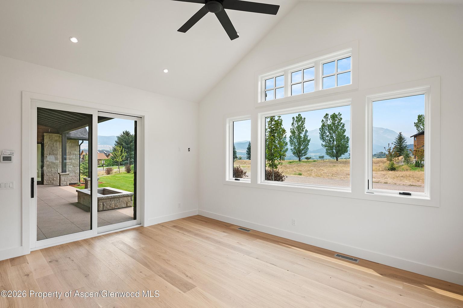 This bright and airy living room features light wood flooring and crisp white walls, creating a clean, modern aesthetic. A large sliding glass door provides seamless access to an outdoor patio, while a series of expansive windows offers panoramic views of the surrounding landscape and mountains. The vaulted ceiling, accented by a dark ceiling fan, enhances the sense of space and openness in this minimalist room.