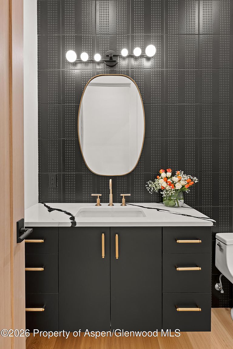 This modern powder room features a striking dark, textured tile wall accented by a sleek, oval-shaped mirror and a contemporary vanity with gold-toned hardware. The vanity is topped with a white stone surface featuring bold dark veining, complemented by a minimalist faucet and a small floral arrangement. The perspective is a straight-on, eye-level shot that highlights the high-contrast design and sophisticated finishes of the space.